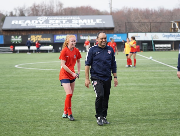 A group of people are on a soccer field, with some wearing sports attire. In the foreground, a person in red sports clothing is walking alongside someone in a dark tracksuit. Other people are visible in the background, some wearing yellow jerseys. A building with advertising banners, including a prominent sign reading 'Ready, Set, Smile,' is in the background.